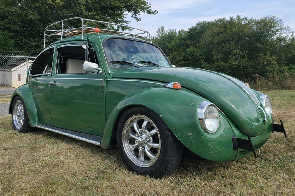Green Volkswagen Super Beetle with roof rack parked on grass near trees and a fence