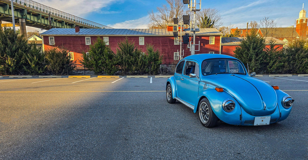 Bright blue Volkswagen Super Beetle parked in front of a red building and bridge.